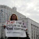 На фото и фоне правительства Москвы стоит женщина с плакатом, на котором написано: 27 лет стою в очереди на получение квартиры, живу в коммуналке.