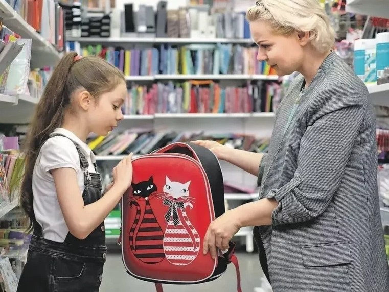 На фото мама собирает дочку в школу покупая рюкзак и канцелярские товары.