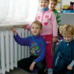 На фото дети в детском саду греются возле батареи центрального отопления.