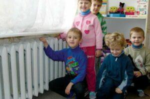 На фото дети в детском саду греются возле батареи центрального отопления.